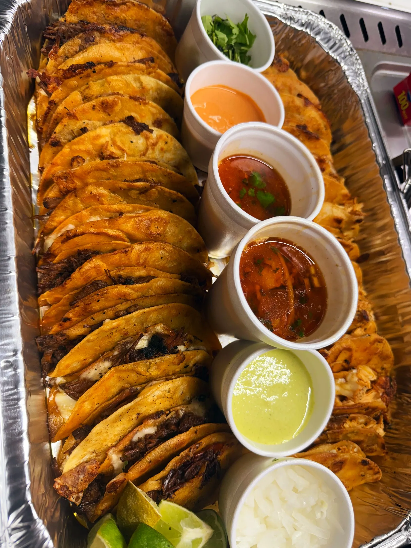 A catering pan packed with birria tacos, cups of consomé and salsas, onion and lime