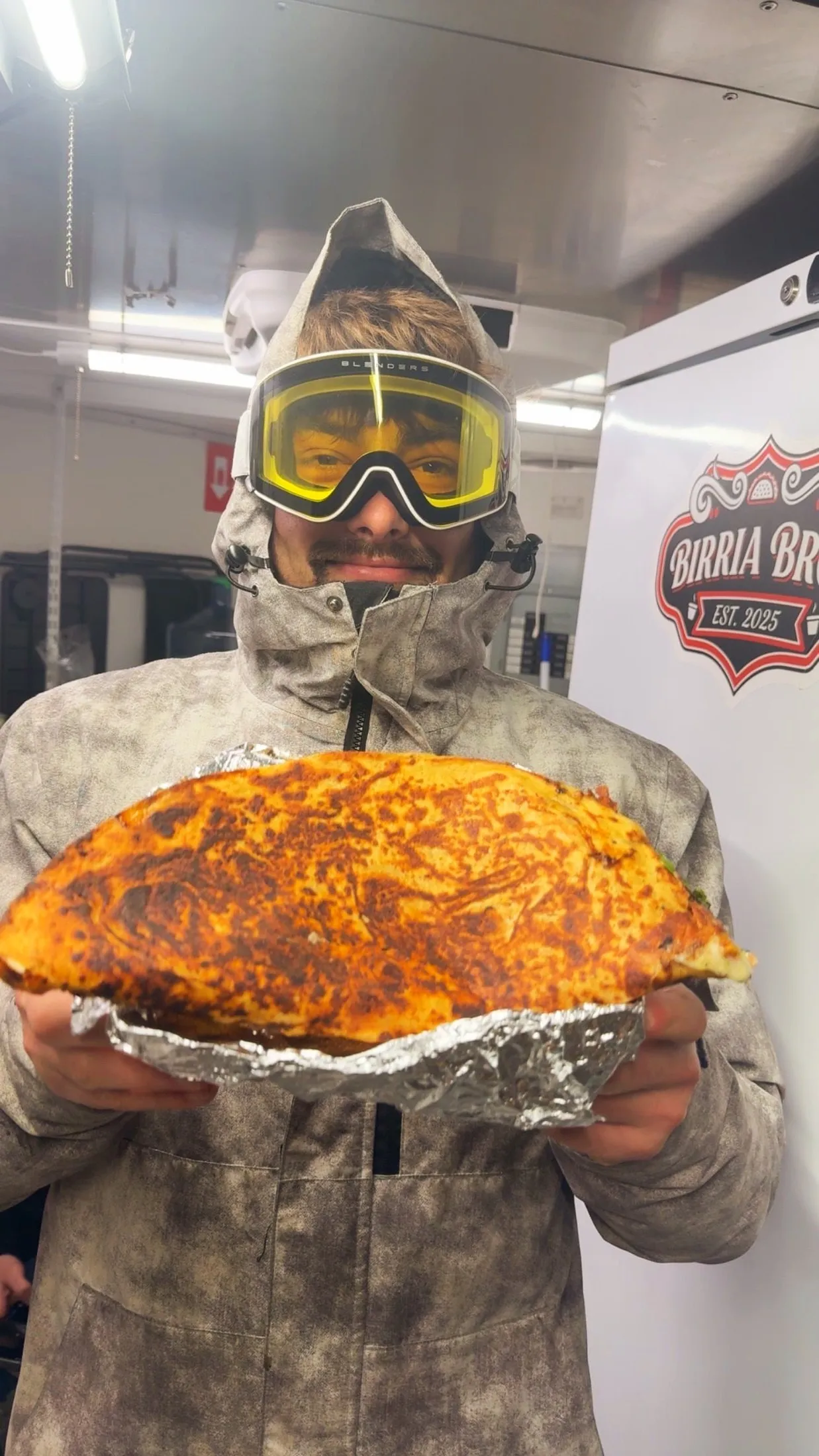 A guy in ski goggles and a winter jacket holding a giant quesadilla inside the truck