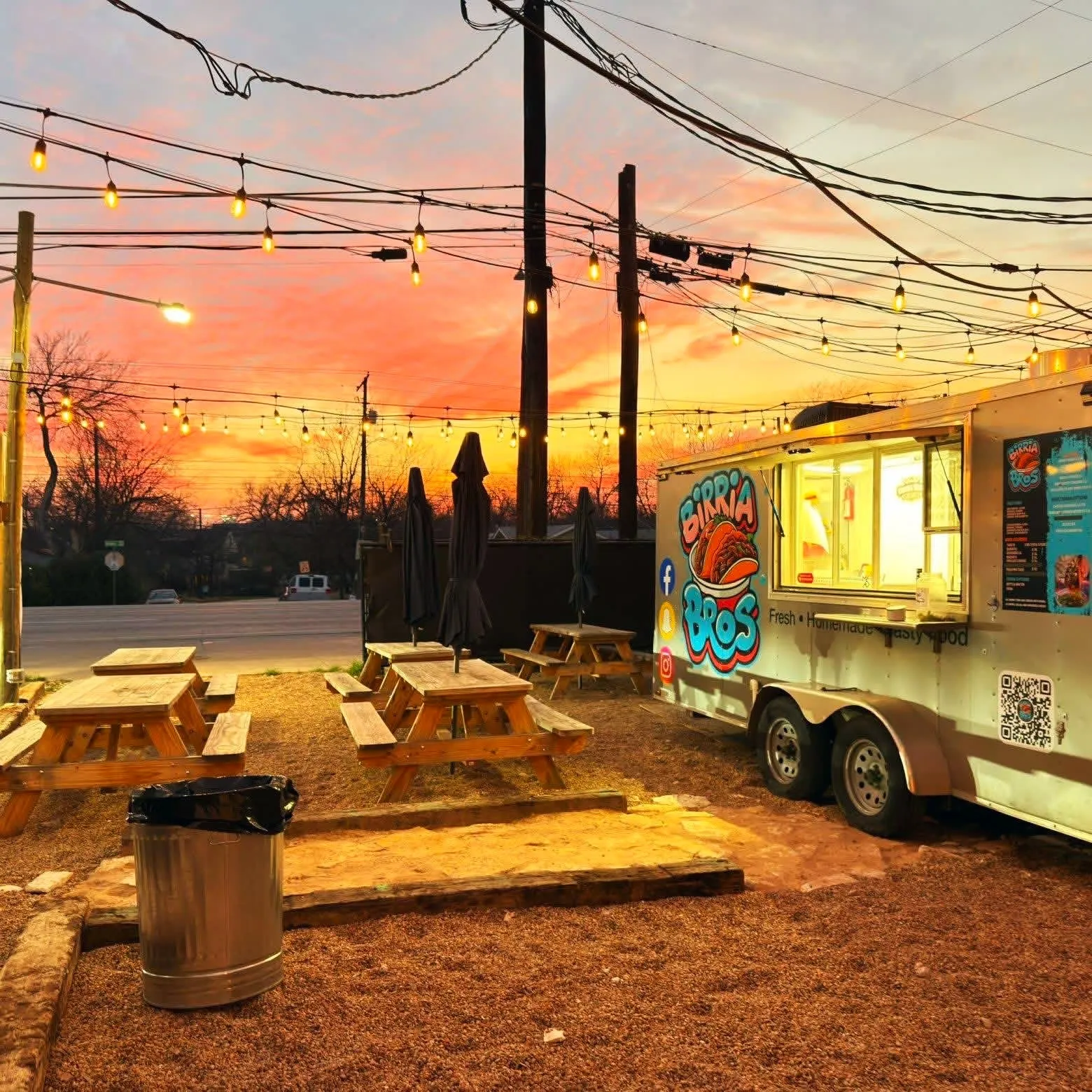 The Birria Bros truck at a patio with picnic tables and string lights at sunset